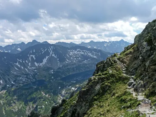 Kamienista ścieżka wije się wzdłuż skalistego grzbietu w Tatrach Wysokich. W oddali widać ośnieżone szczyty i jezioro.