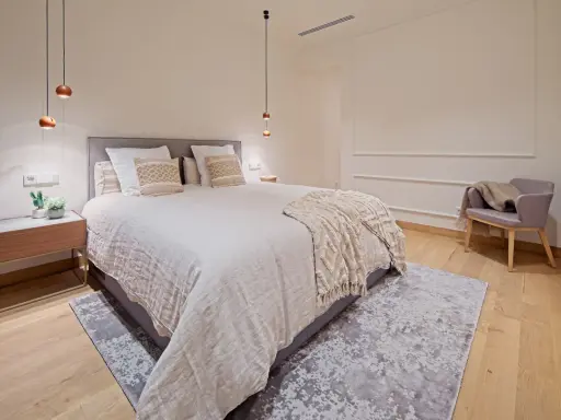 Dormitorios relajantes con cama amplia, manta texturizada, sillón acogedor y luces colgantes.