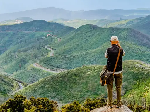 Mann mit Kamera genießt die Aussicht auf kurvige Straße in grünen Bergen. Perfektes Ziel für Wandern in Spanien.