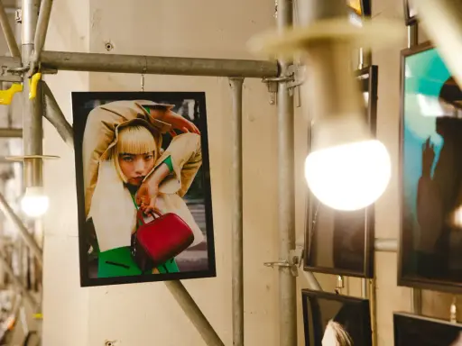 Plakat z kobietą w beżowym płaszczu i zielonym swetrze, trzymającą czerwoną torebkę. Stylizacja przypomina modę z **Pezet Tokyo**.