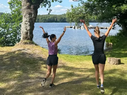 Två kvinnor hoppar glatt vid en sjö, redo att springa en mil om dagen i den vackra svenska naturen.