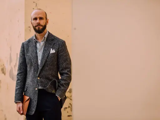 Man in a textured grey jacket and black pants, holding a tan envelope clutch.