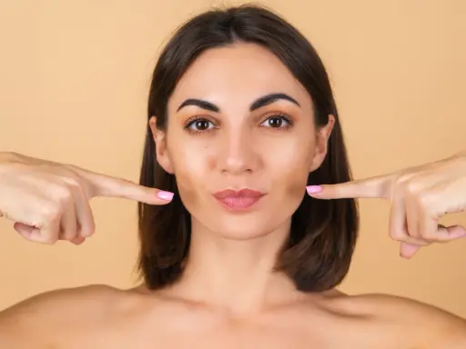 Doigts pointés vers les joues, une femme montre comment repulper son visage pour un look plus jeune et plein.