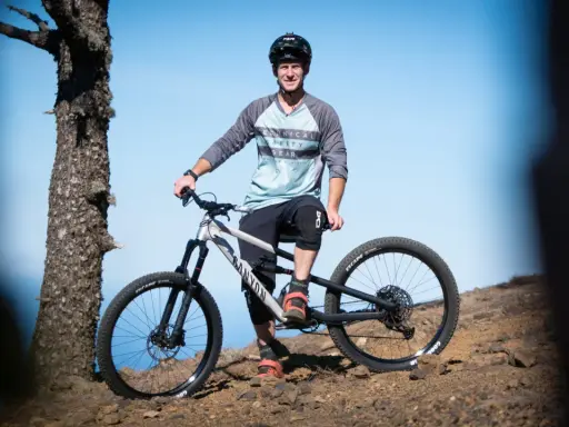 Sam Pilgrim, bereit für die nächste Abfahrt, posiert mit seinem Mountainbike auf einem felsigen Hang vor blauem Himmel.