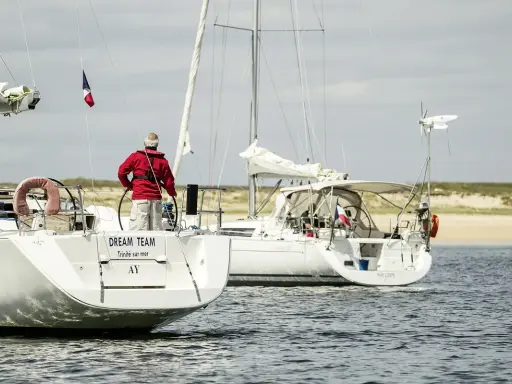 Un homme en gilet rouge tient la barre d'un voilier, le pont est bien mouillé. Un autre voilier suit, naviguant sur une mer calme.