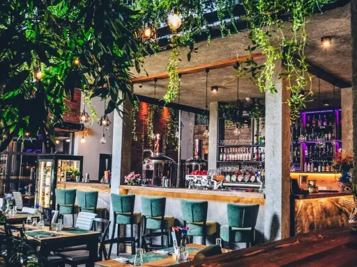 Wnętrze hotelu z browarem: bar z zielonymi hokerami, miedziany destylator, półki z butelkami i mnóstwo zieleni.
