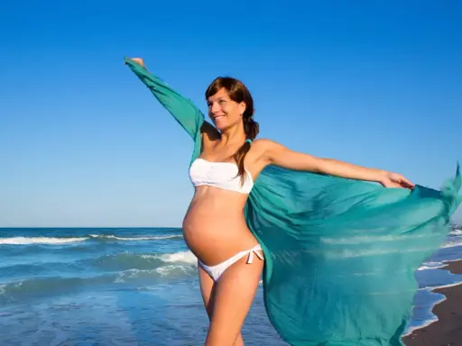 Donna incinta in bikini bianco sulla spiaggia, con un velo turchese che svolazza al vento. Il mare sullo sfondo.