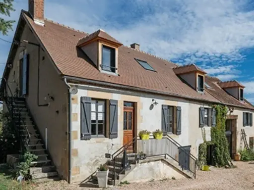 Une maison de campagne simple avec des volets gris, une cheminée et des escaliers extérieurs sous un ciel bleu.