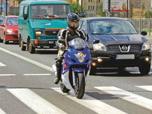 Motocyklista w kasku i kombinezonie na pasach. Obok samochody i furgonetka. Pamiętaj o przeglądzie, bo jaka kara za brak przeglądu może być dotkliwa.