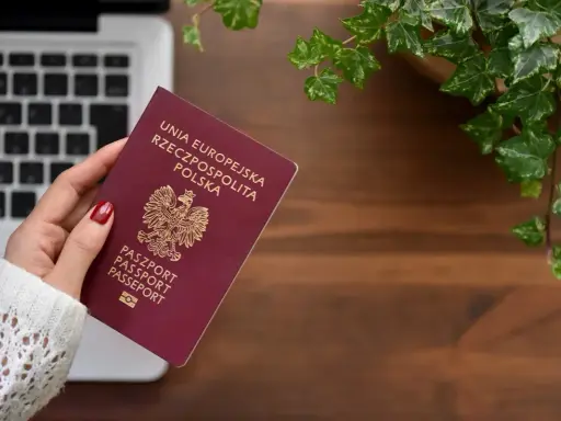 Dłoń trzyma polski paszport na tle laptopa i roślin.