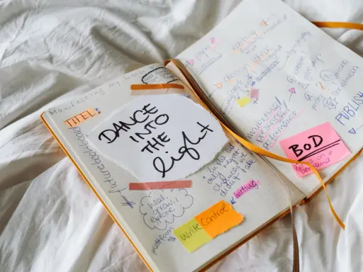 Carnet ouvert avec des notes manuscrites sur la création d'un livre, incluant "Dance into the Light" et des idées de marketing. Un nom d'auteur connu pourrait être inspiré par ces notes.