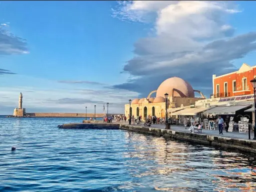 Il porto di Chania con il suo faro iconico e la moschea ottomana, bagnato da un mare blu intenso sotto un cielo nuvoloso.