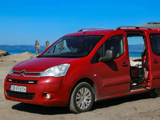 Czerwony Citroen Berlingo 1.6 HDI na plaży, z otwartymi drzwiami i bagażnikiem, na tle morza i palmy.