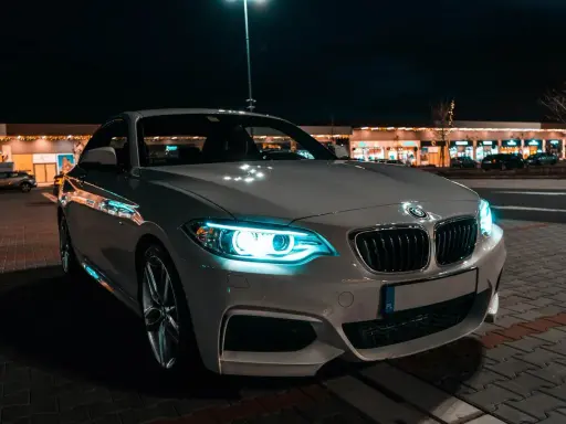 Białe BMW z włączonymi światłami adaptacyjnymi błyszczy nocą przed sklepami.