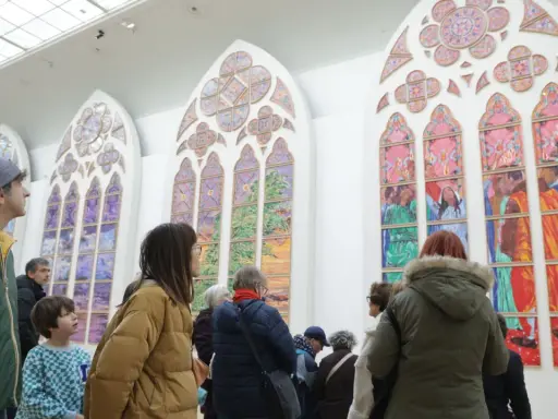 Des visiteurs contemplent de grands vitraux contemporains aux couleurs vives, représentant des scènes et des paysages.