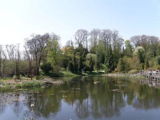 Spokojny staw w Bolestraszycach, otoczony drzewami i zielenią. Widoczna drewniana kładka z ludźmi, idealne miejsce na relaks i podziwianie atrakcji.