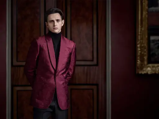 A man in a patterned burgundy jacket and black turtleneck, perfect for cocktail party attire for men, stands in a dimly lit room with ornate wooden doors.