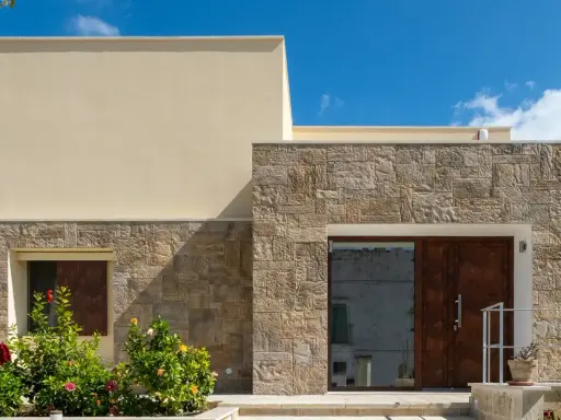 Moderne facciate in pietra grezza si alternano a intonaco chiaro, creando un contrasto materico. Un ingresso con porta in legno scuro e un giardino fiorito.