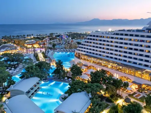 Luksusowy hotel Titanic Deluxe Lara z basenami i parkiem wodnym, oświetlony wieczorem, z widokiem na morze i góry.