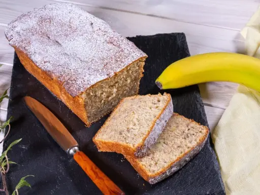 Ein **super saftiger Bananenkuchen**, bestäubt mit Puderzucker, liegt auf einem schwarzen Stein neben zwei Scheiben. Eine Banane und ein Messer liegen daneben.