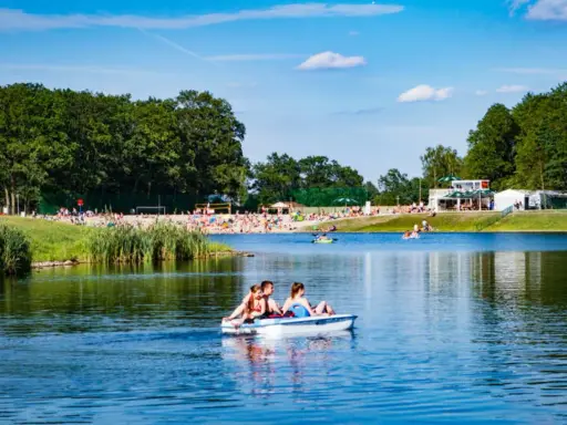 Rodzina pływa rowerkiem wodnym po jeziorze na Dolnym Śląsku, w tle plaża pełna ludzi i zielone drzewa.