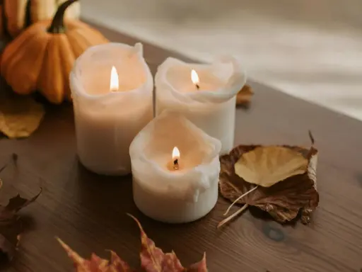 Candele accese su un tavolo di legno, con zucche e foglie autunnali. Un'ispirazione per chi vuole imparare come fare candele in casa.