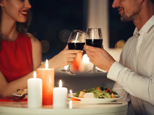 Romantyczna kolacja przy świecach. Para wznosi toast kieliszkami czerwonego wina, uśmiechając się do siebie.