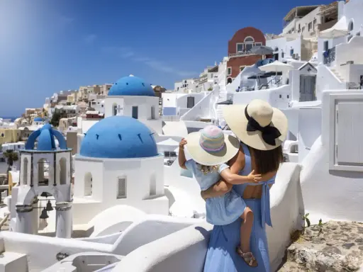 Una madre abbraccia la sua bambina, ammirando il panorama di Santorini, Grecia. Le cupole blu brillano sotto il sole, mentre le case bianche si aggrappano alla collina.