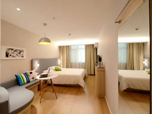 Habitación estrecha y alargada con cama, sofá curvo, mesa con portátil y espejo de cuerpo entero.