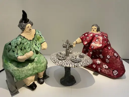 Dos figuras voluminosas, al estilo de Niki de Saint Phalle, sentadas a una mesa de té. Una viste de verde, la otra de rojo.