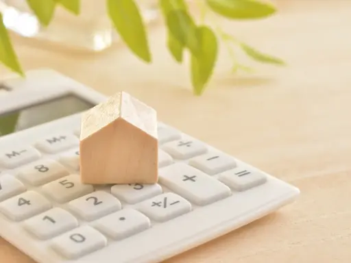 Maison en bois sur une calculatrice blanche, symbolisant le calcul du prix du **pel plafond**. Des feuilles vertes floues en arrière-plan.