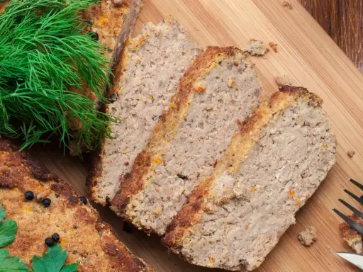 Pokrojony pasztet drobiowy z wątróbką, ozdobiony koperkiem i natką pietruszki, czeka na desce do krojenia.