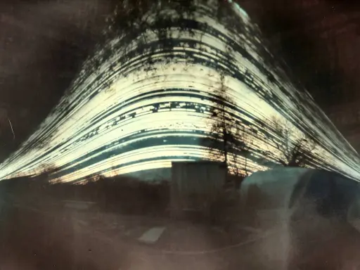 Paesaggio distorto con alberi e strutture, catturato con un **foro stenopeico**. Le strisce luminose suggeriscono un lungo tempo di esposizione.