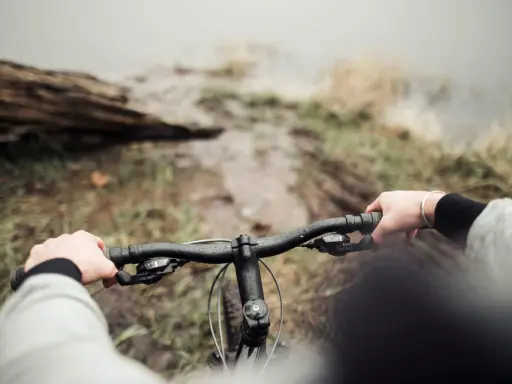 Ręce na kierownicy roweru, błotnista ścieżka i widok na wodę. To właśnie dlatego warto jeździć rowerem – bliskość natury i wolność.