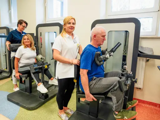Rehabilitacja w Busko Zdrój: pan ćwiczy na siłowni pod okiem fizjoterapeutki, a pani z pomocą drugiej terapeutki. Sprawdź nasze sanatoria Busko Zdrój promocje!
