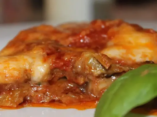 Deliciosa porción de parmigiana di melanzane, con capas de berenjena, salsa de tomate y queso fundido, adornada con una hoja de albahaca fresca.