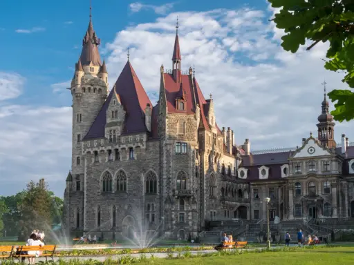 Imponujący zamek a pałac z kamienia, z wieżami i czerwonymi dachami, otoczony zielonym parkiem, gdzie odpoczywają ludzie.