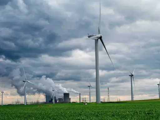 Windräder auf grüner Wiese unter dramatischem Himmel, im Hintergrund rauchende Industrieanlagen. Ein Symbol für Investitionen in erneuerbare Energien in Deutschland.