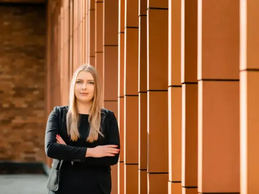 Kobieta w czarnym garniturze stoi na tle ceglanej ściany i pomarańczowych pionowych elementów.