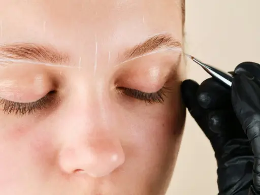 Une main gantée dessine des lignes blanches sur un sourcil pour un microblading poil à poil.