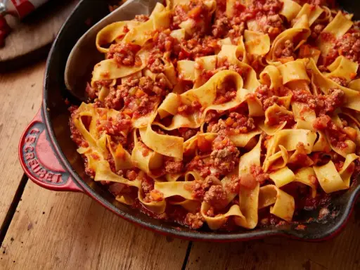 En stor portion pasta bolognese med breda bandnudlar och en mustig köttfärssås i en röd gjutjärnspanna.