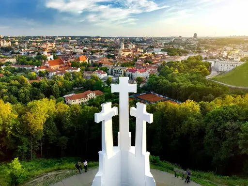 Wzgórze Trzech Krzyży w Wilnie z widokiem na miasto. Trzy białe krzyże górują nad zielonymi drzewami i zabytkową architekturą.
