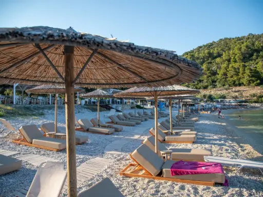 Alonissos spiagge: lettini e ombrelloni di paglia su una spiaggia di ciottoli, con il mare cristallino e la collina verde sullo sfondo.