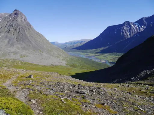 Vacker vy över Sareks nationalpark med Rapadalen i fjärran. En flod slingrar sig genom en grön dal omgiven av höga, steniga berg under en klarblå himmel.