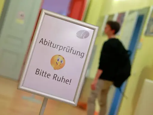 Schild mit der Bezeichnung "Abiturprüfung. Bitte Ruhe!" und einem Emoji, das zum Schweigen auffordert. Ein Schüler geht im Hintergrund vorbei.