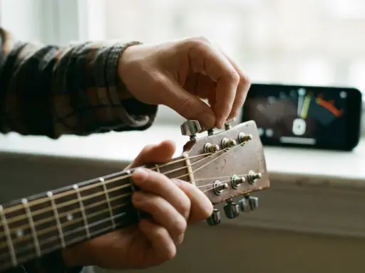 Main d'un musicien ajustant une guitare accordée, avec un accordeur sur un téléphone en arrière-plan.