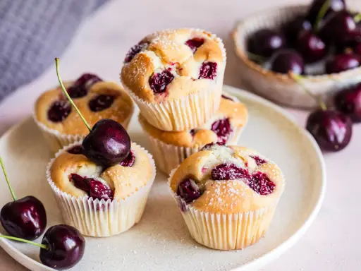 Saftige Muffins mit Sauerkirschen, bestäubt mit Puderzucker. Frische Kirschen liegen daneben und auf einem Muffin.