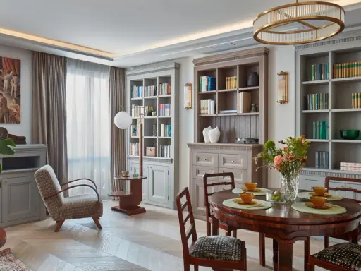 Elegancki salon w stylu art deco z dwoma regałami pełnymi książek, okrągłym stołem i wygodnym fotelem.