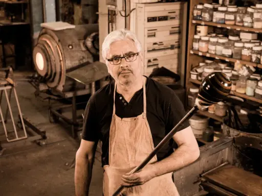 Un artisan verrier expérimenté, tablier en cuir, tient sa canne à souffler devant son four incandescent.