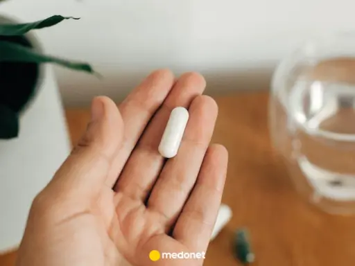Dłoń trzyma białą kapsułkę. Obok szklanka wody i roślina. Uważaj na przedawkowanie magnezu.